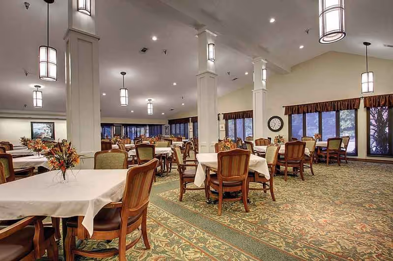 Spacious dining room with multiple tables draped in white cloths, wooden chairs, pendant lights, and large windows.