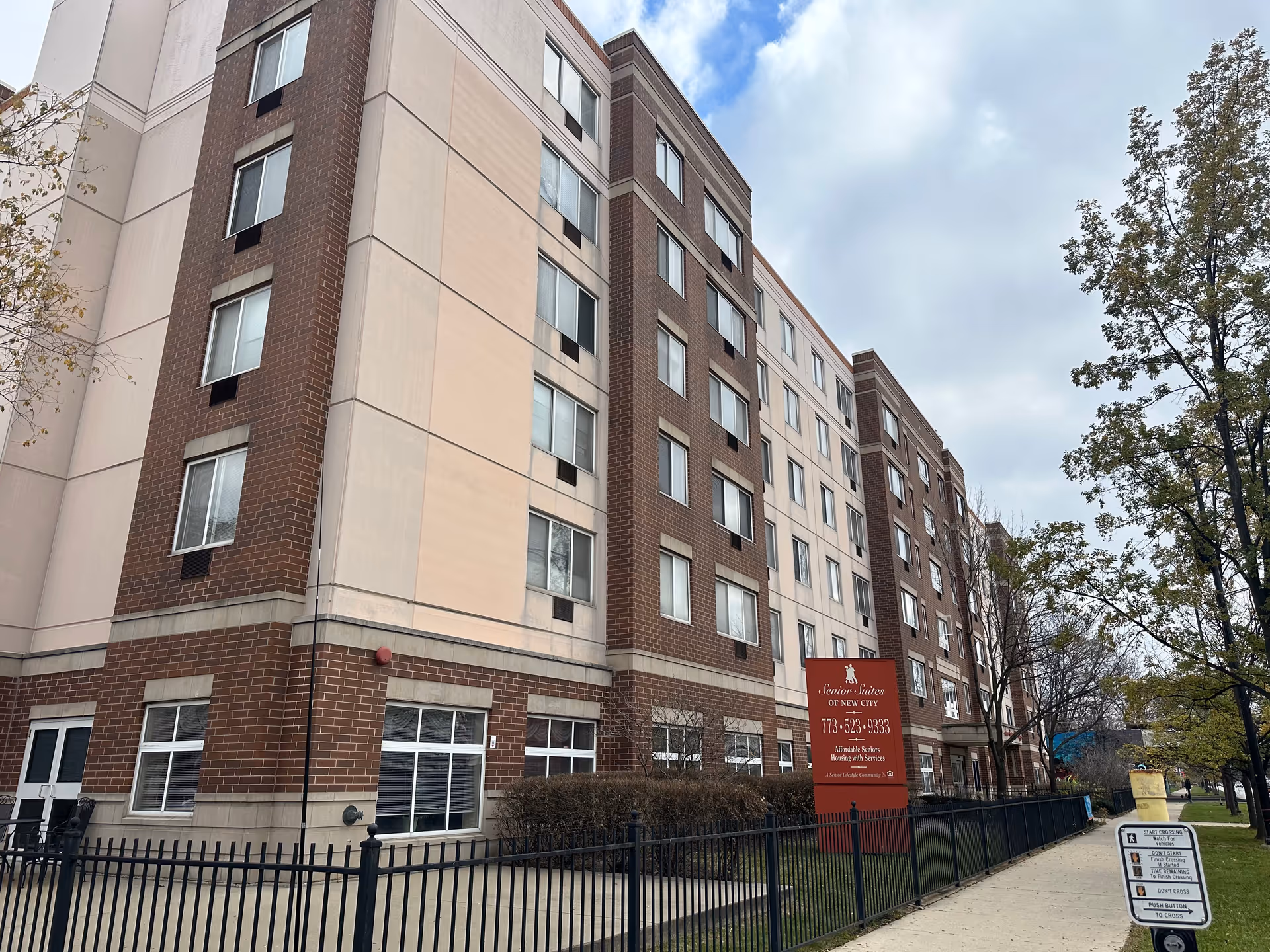 Wide exterior view of the multi-story Senior Suites of New City building with brick and light-colored panels, a fenced sidewalk, trees, and a red identification sign.