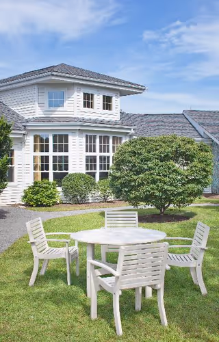 Outdoor seating area with a round white table and four white chairs on a grassy lawn in front of a white building with large windows and a gray roof under a partly cloudy sky.