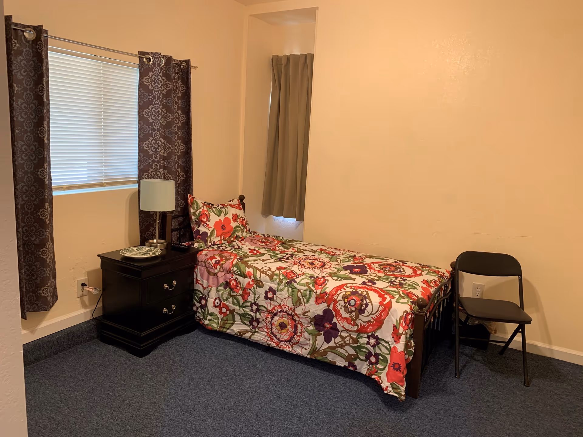 Small bedroom with a single bed in a floral comforter, a dark nightstand with a lamp by a curtained window, and a folding chair.