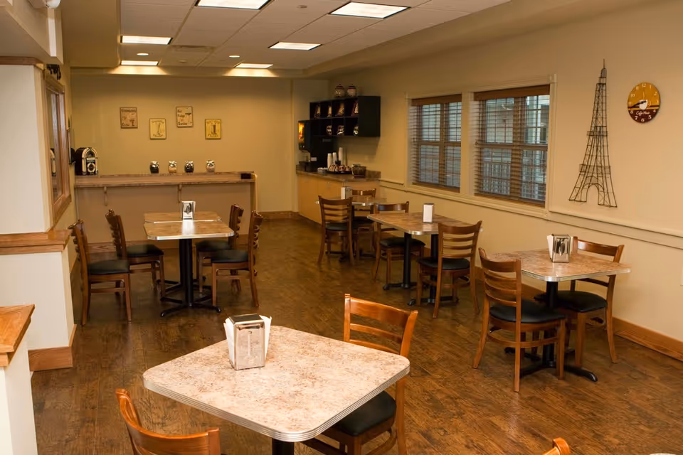 A small dining area with several square tables and wooden chairs arranged neatly. The room has wooden flooring and beige walls with windows on one side. There is a counter at the back with coffee dispensers and cups, and wall decorations including a wireframe Eiffel Tower and a round clock.