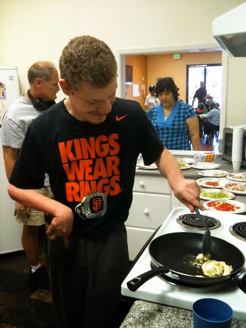 A man cooks at a stove in a kitchen while others chat in background.