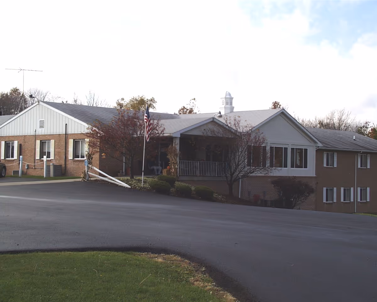 Exterior view of a single-story assisted living facility building with a sloped roof, several windows, a porch area with a railing, an American flag on a flagpole, and some small trees and bushes in front. The driveway is paved and curves in front of the building.