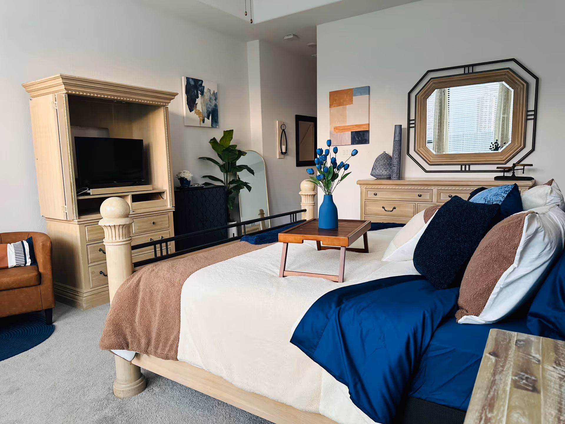 Bright bedroom featuring a large bed with blue and tan bedding, dresser, mirror, and TV unit.