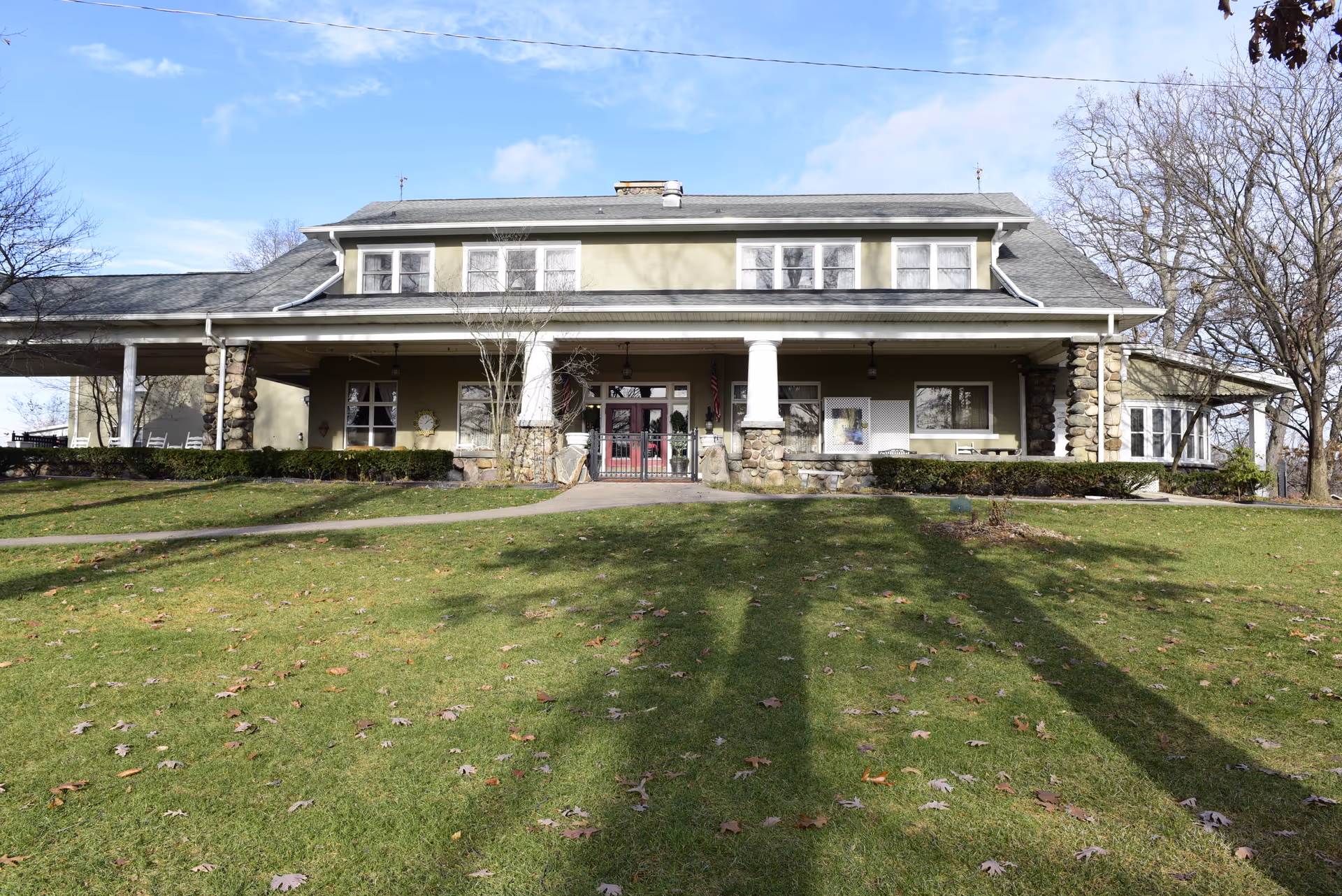 Front exterior view of a large two-story building with a covered porch supported by stone pillars, surrounded by a grassy lawn with some trees and a clear blue sky.