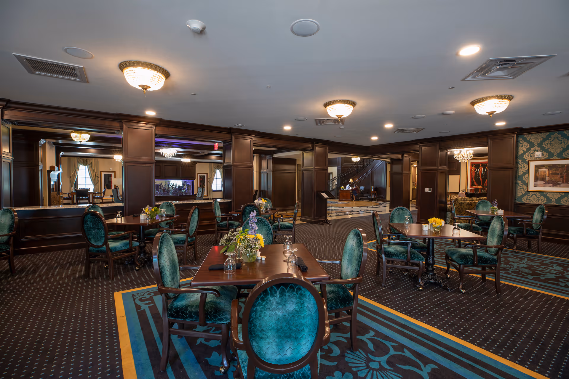 Elegant senior living dining room with dark wood panels, green upholstered chairs, round chandeliers, and flowers.