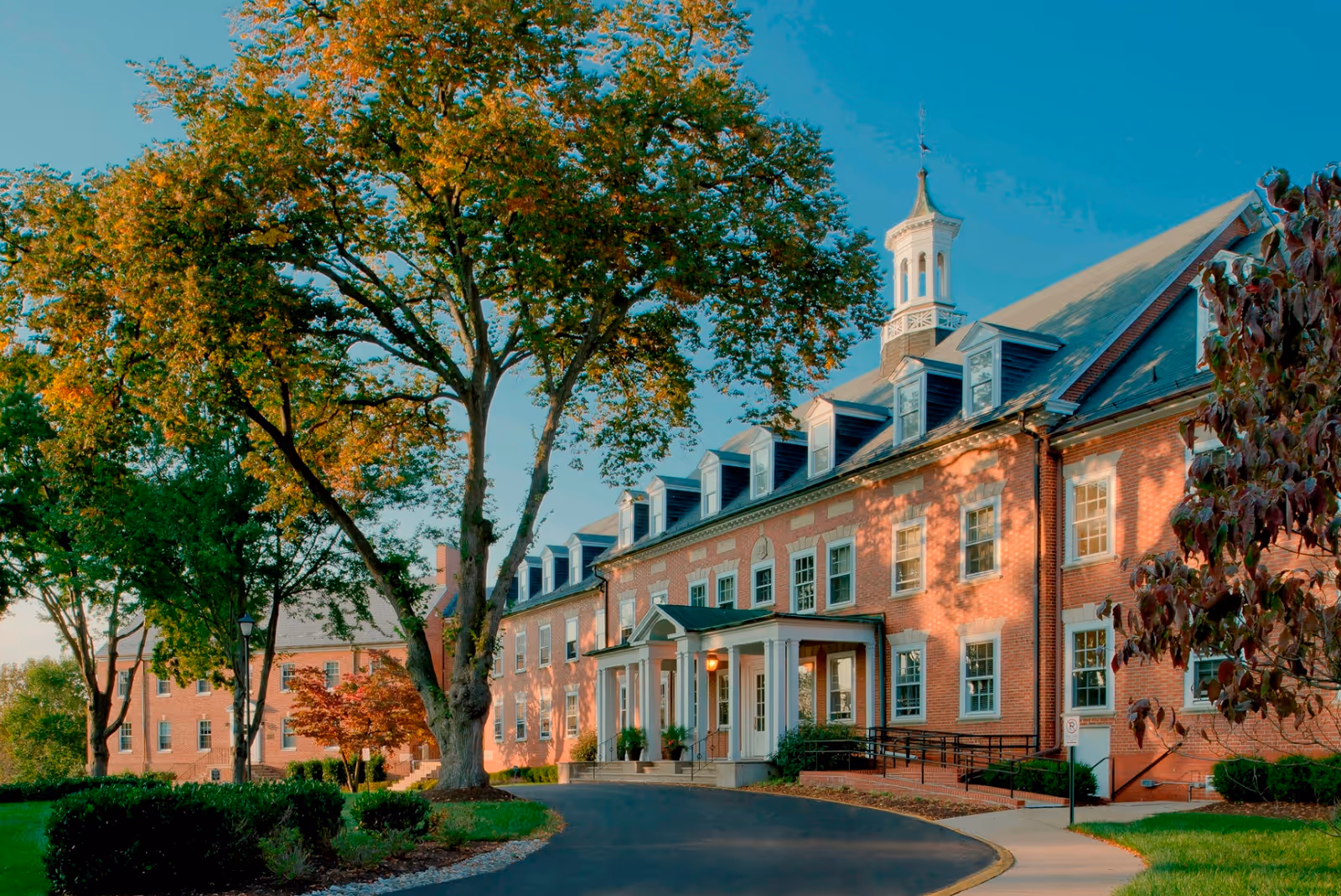 Exterior view of a large brick building with multiple windows and a cupola on the roof, surrounded by trees with autumn foliage and a curved driveway leading to the entrance.