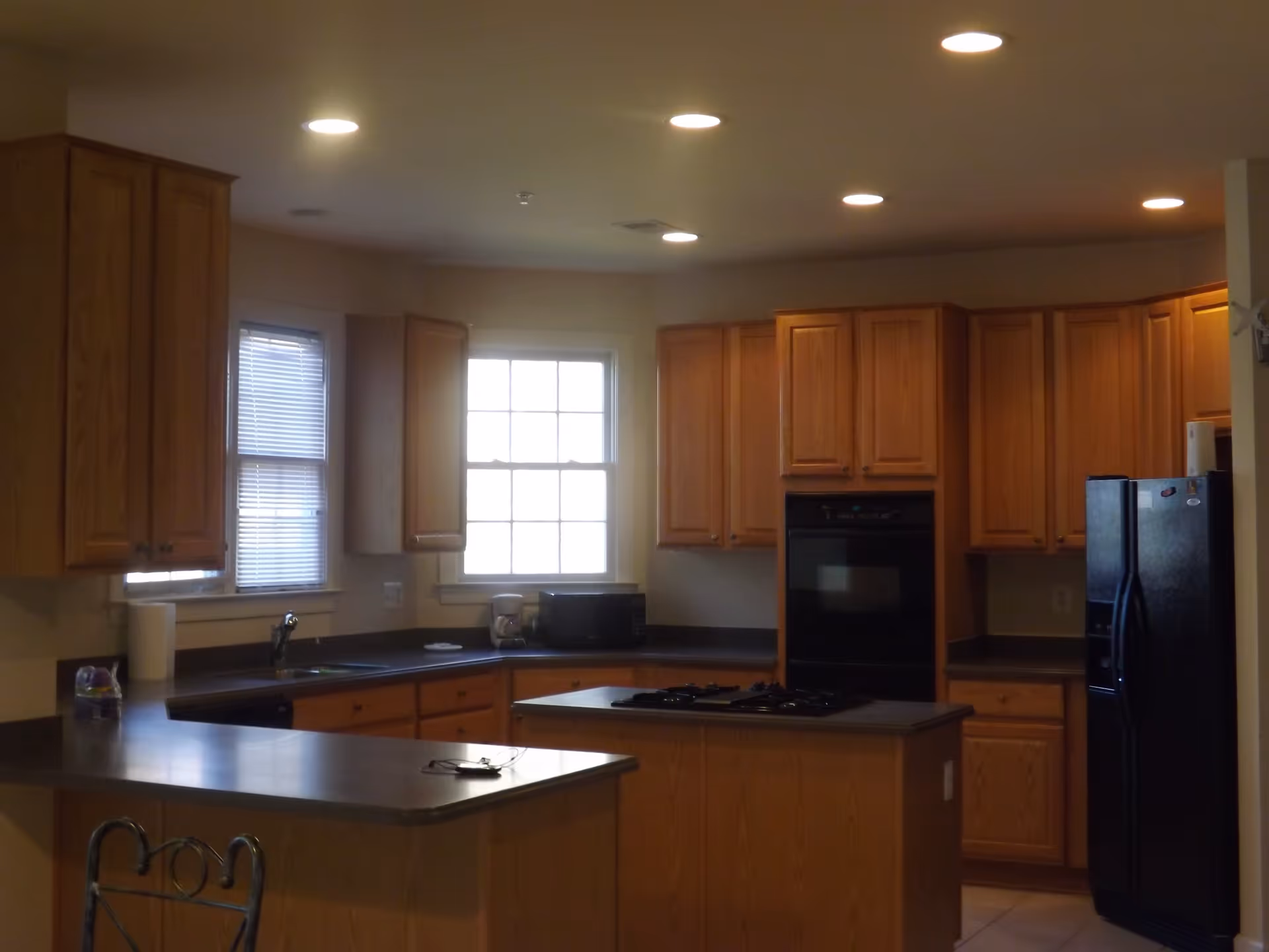 Interior view of a kitchen with wooden cabinets, a black refrigerator, a built-in oven, a microwave, a coffee maker, and a kitchen island with a stovetop. There are two windows with blinds and recessed ceiling lights.