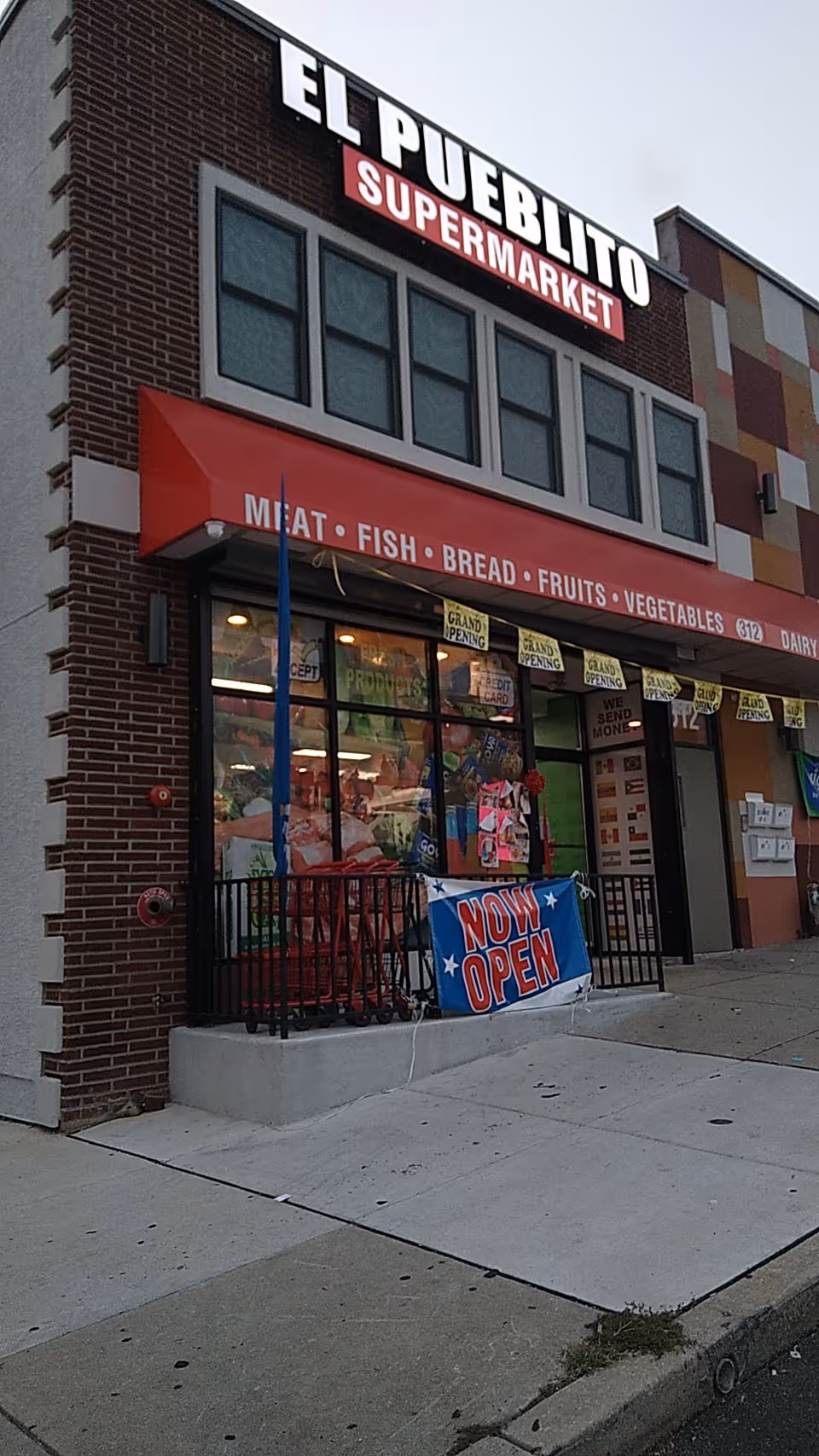 Exterior view of a supermarket storefront with red awning and 'NOW OPEN' banner