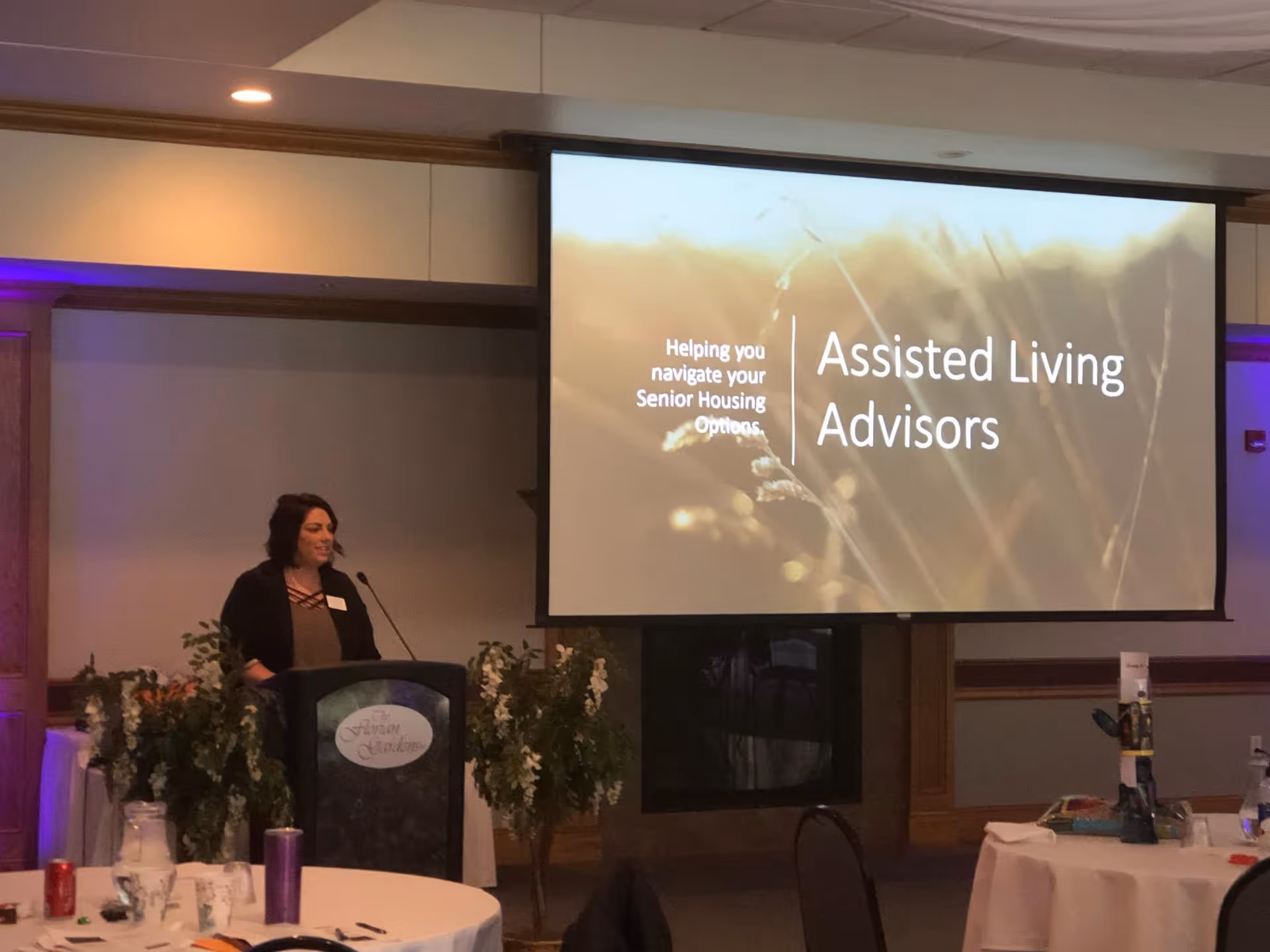 A speaker at a podium beside a large screen displaying Assisted Living Advisors.