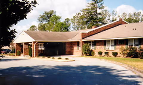Exterior view of a single-story brick building with a sloped roof, surrounded by trees and a paved driveway in front.