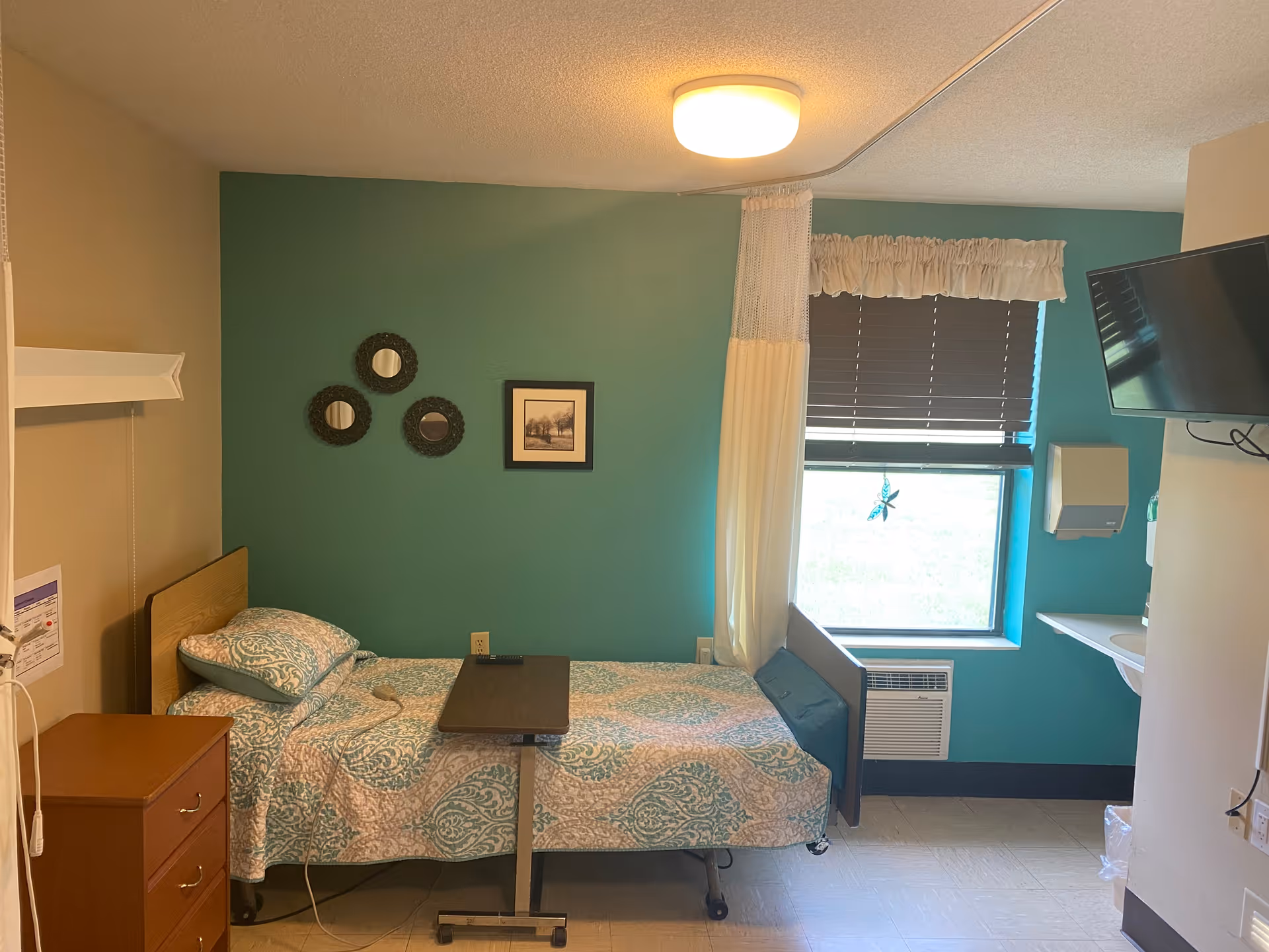 Small single-occupancy senior care bedroom with a hospital-style bed, overbed table, teal accent wall, window, wall-mounted TV and dresser.