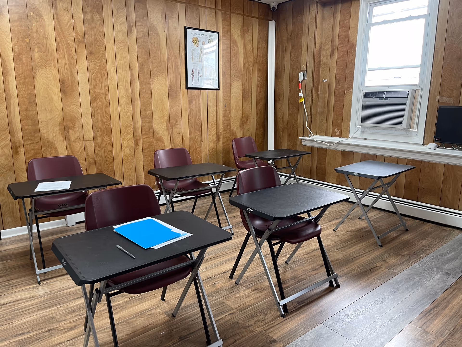 A classroom-style room with folding desks, burgundy chairs, wooden paneled walls, and a window air conditioner.