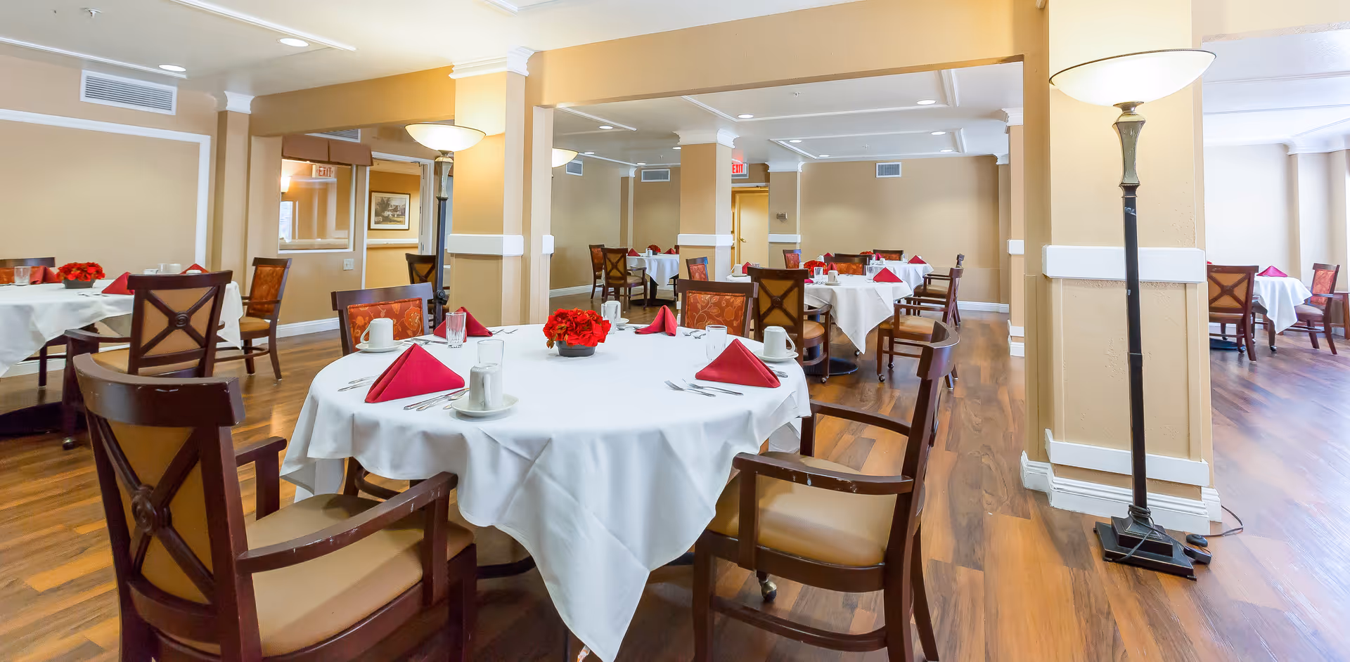 A spacious dining room with round tables, white tablecloths, red napkins, and wooden chairs.