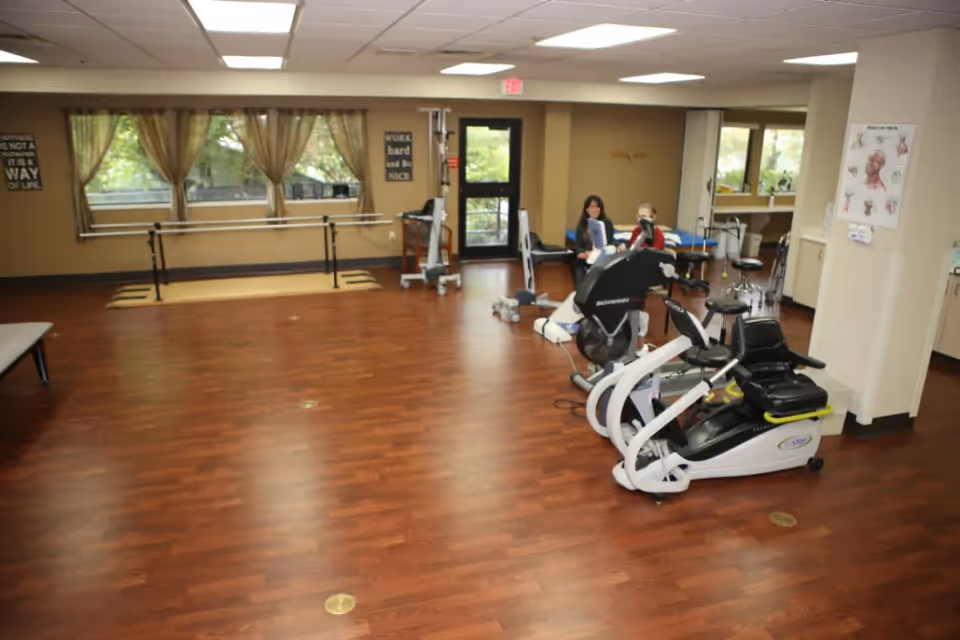 A spacious room with wooden flooring featuring exercise equipment including stationary bikes and parallel bars. Two people are seated near the exercise machines. The room has large windows with curtains, motivational wall signs, and medical posters on the walls.