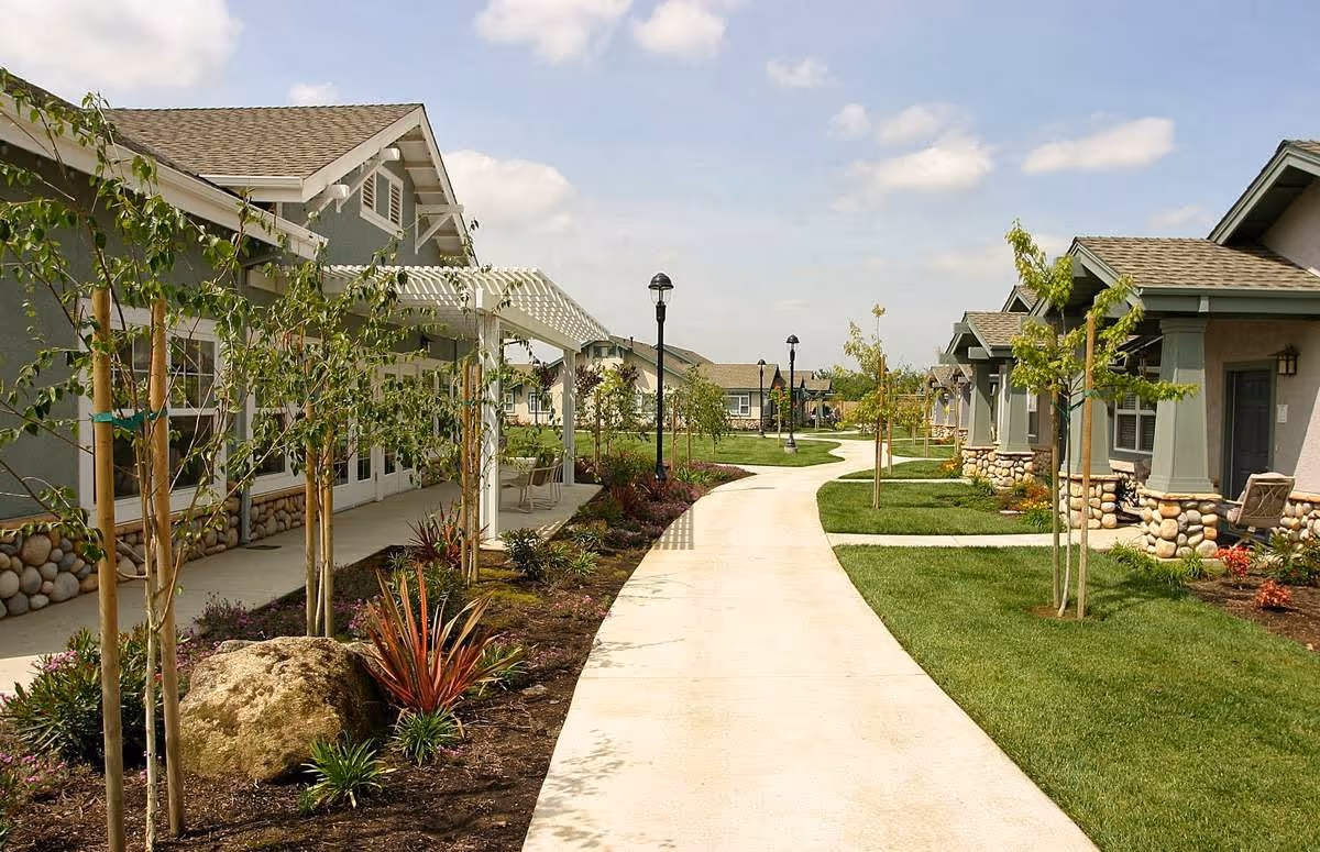 A paved walkway winds through a landscaped outdoor area with small trees, shrubs, and grass. On either side of the path are single-story buildings with stone accents and grayish-green walls, under a partly cloudy sky.
