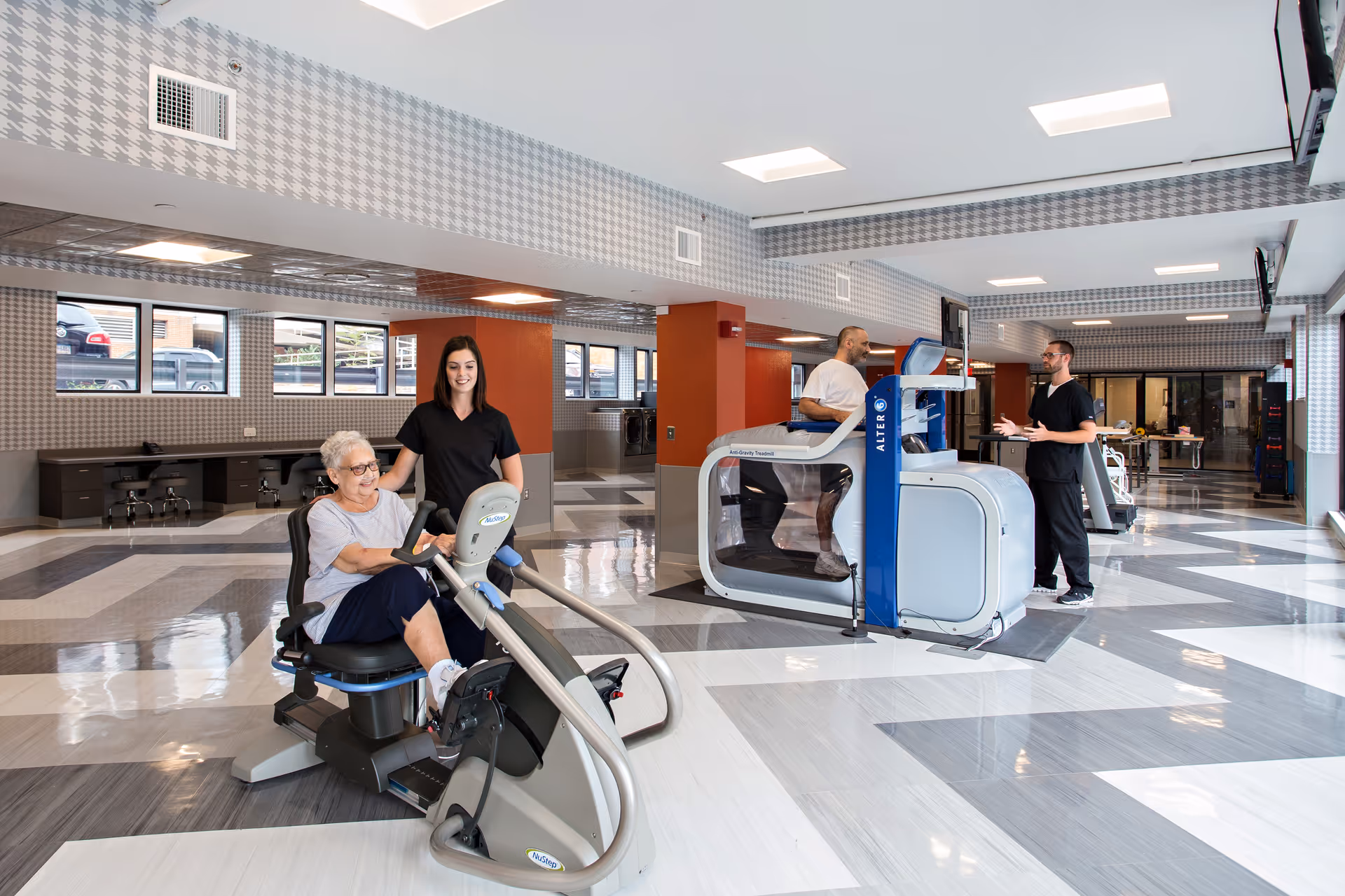 A bright and spacious rehabilitation center room with modern exercise equipment. An elderly woman is using a seated exercise bike while a female staff member stands beside her, smiling. In the background, a man is walking on an AlterG anti-gravity treadmill, assisted by a male staff member. The room has large windows, patterned walls, and a shiny tiled floor.