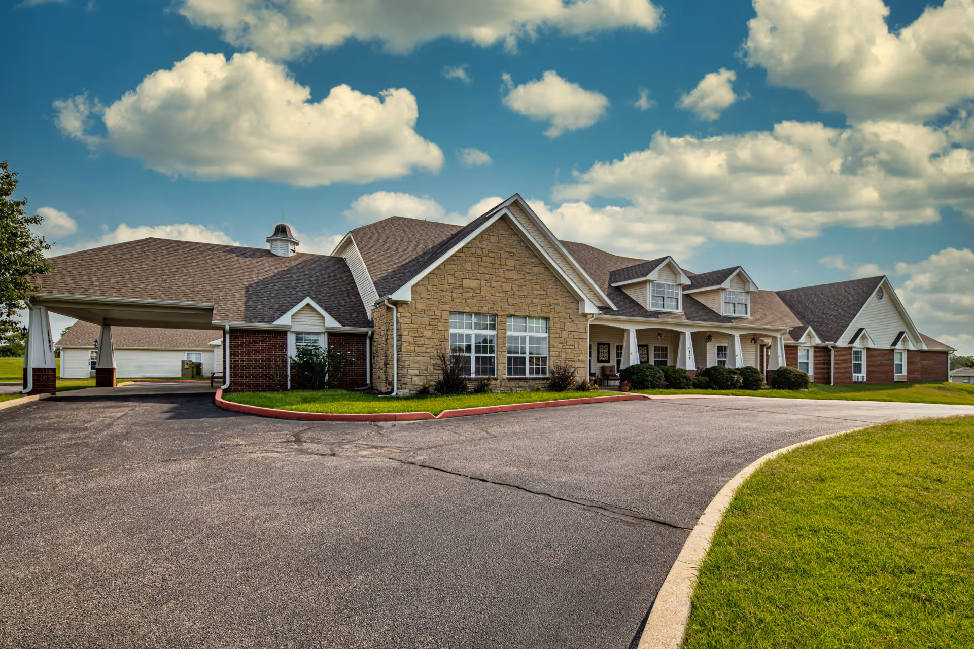 Exterior view of a single-story senior living facility building with a combination of brick and stone facade, multiple windows, a covered entrance, and a paved driveway surrounded by green grass under a partly cloudy sky.