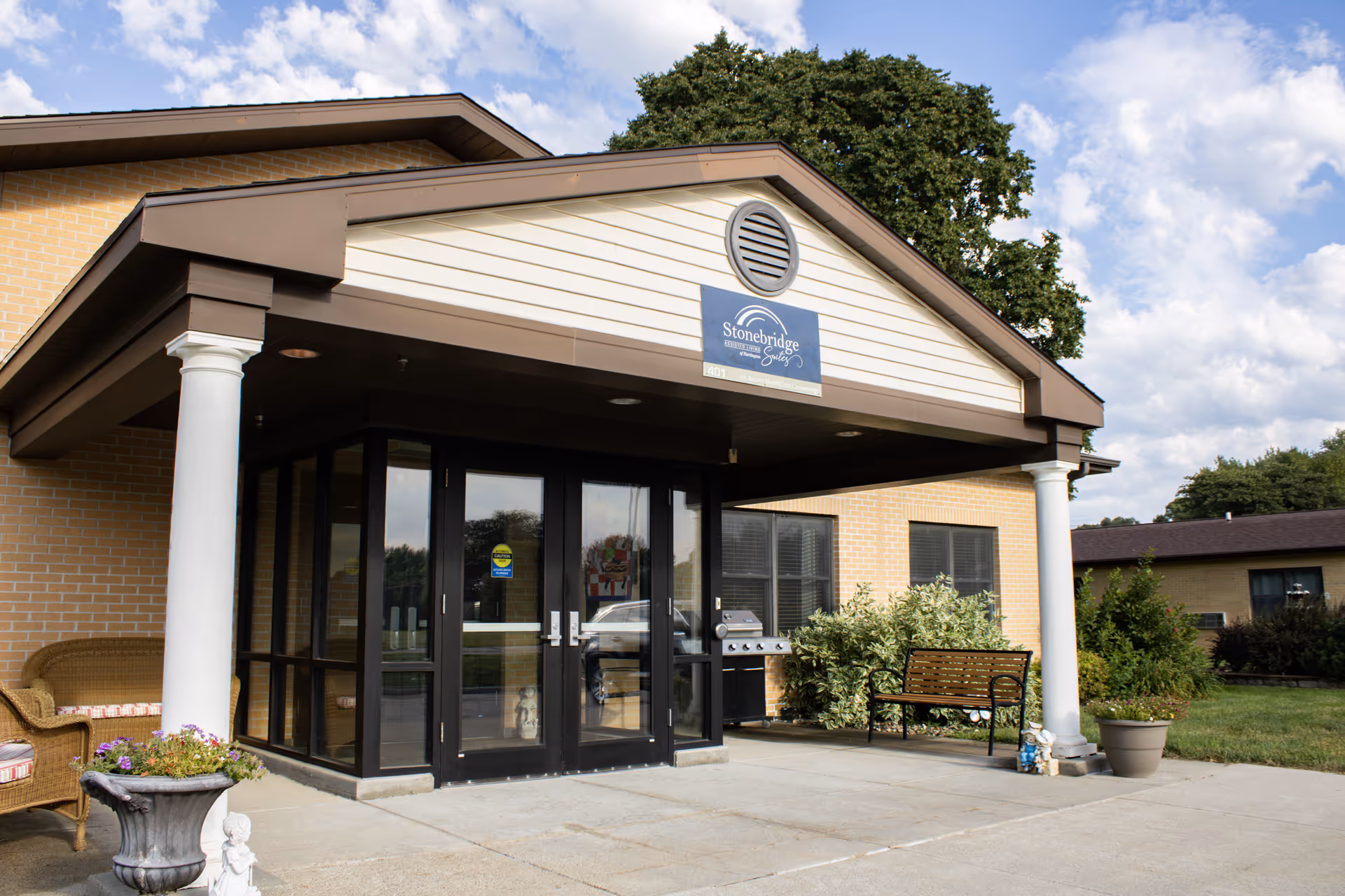 Front entrance of Arbor Care Centers Hartington with glass doors, pillars, bench, and plants.