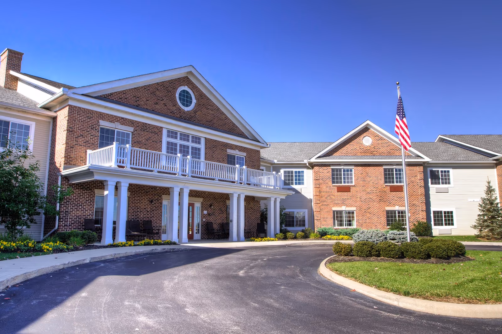 Front entrance of a brick senior living facility with a covered portico, second-floor balcony, and an American flag.