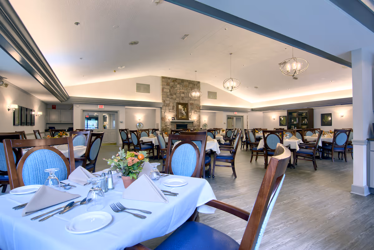 Spacious dining room with set tables, wooden chairs, chandeliers, and a stone fireplace in a senior living community.