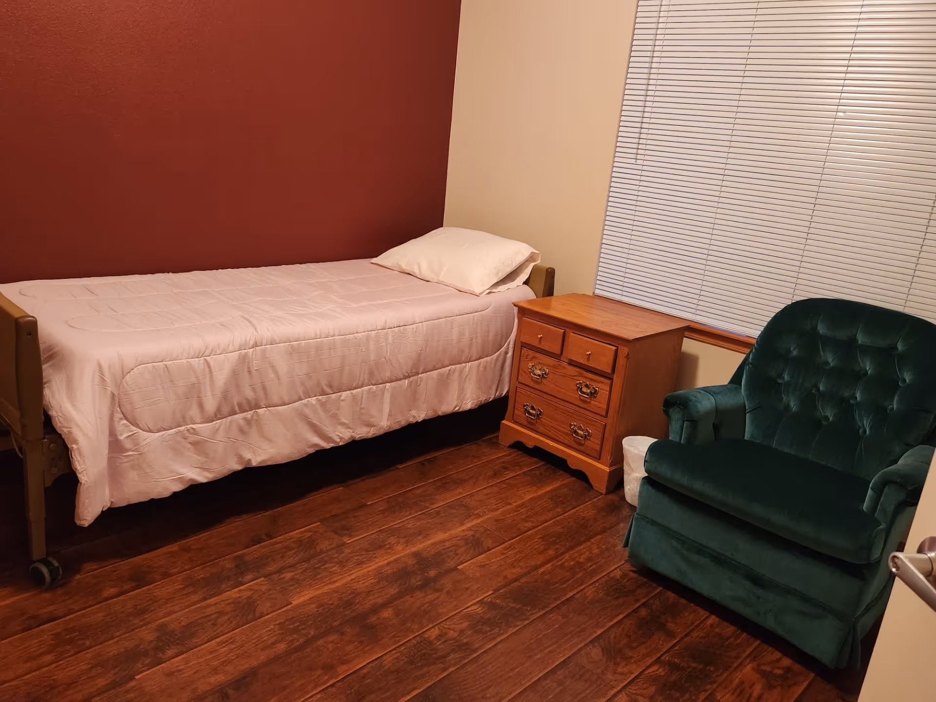 A small bedroom with a single bed covered with a light pink comforter and a white pillow. Next to the bed is a wooden nightstand with drawers, and beside it is a green upholstered armchair. The room has wooden flooring, a maroon accent wall behind the bed, and a window with closed blinds.