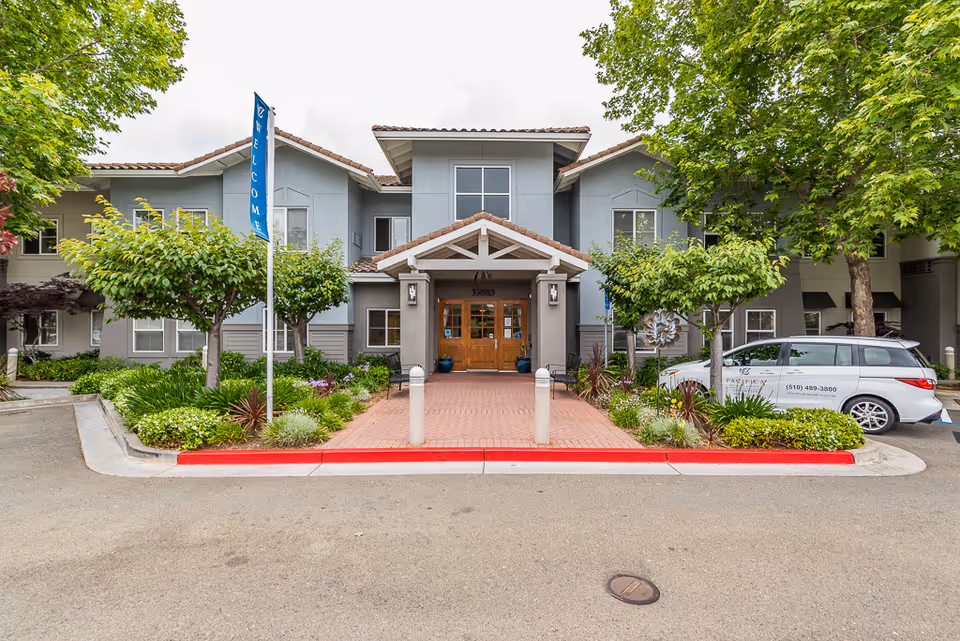 Front entrance of Pacifica Senior Living Union City with a covered portico, landscaped walkway, trees, and a facility van parked to the right.