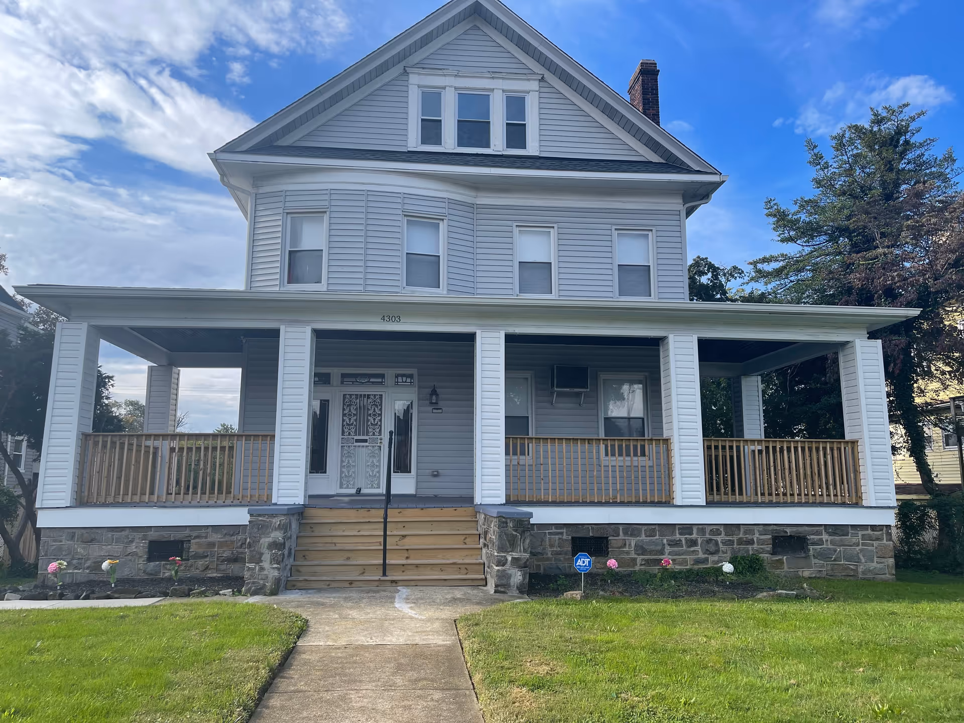 Front view of a two-story light-gray house with a wide covered porch, wooden steps, and a lawn.