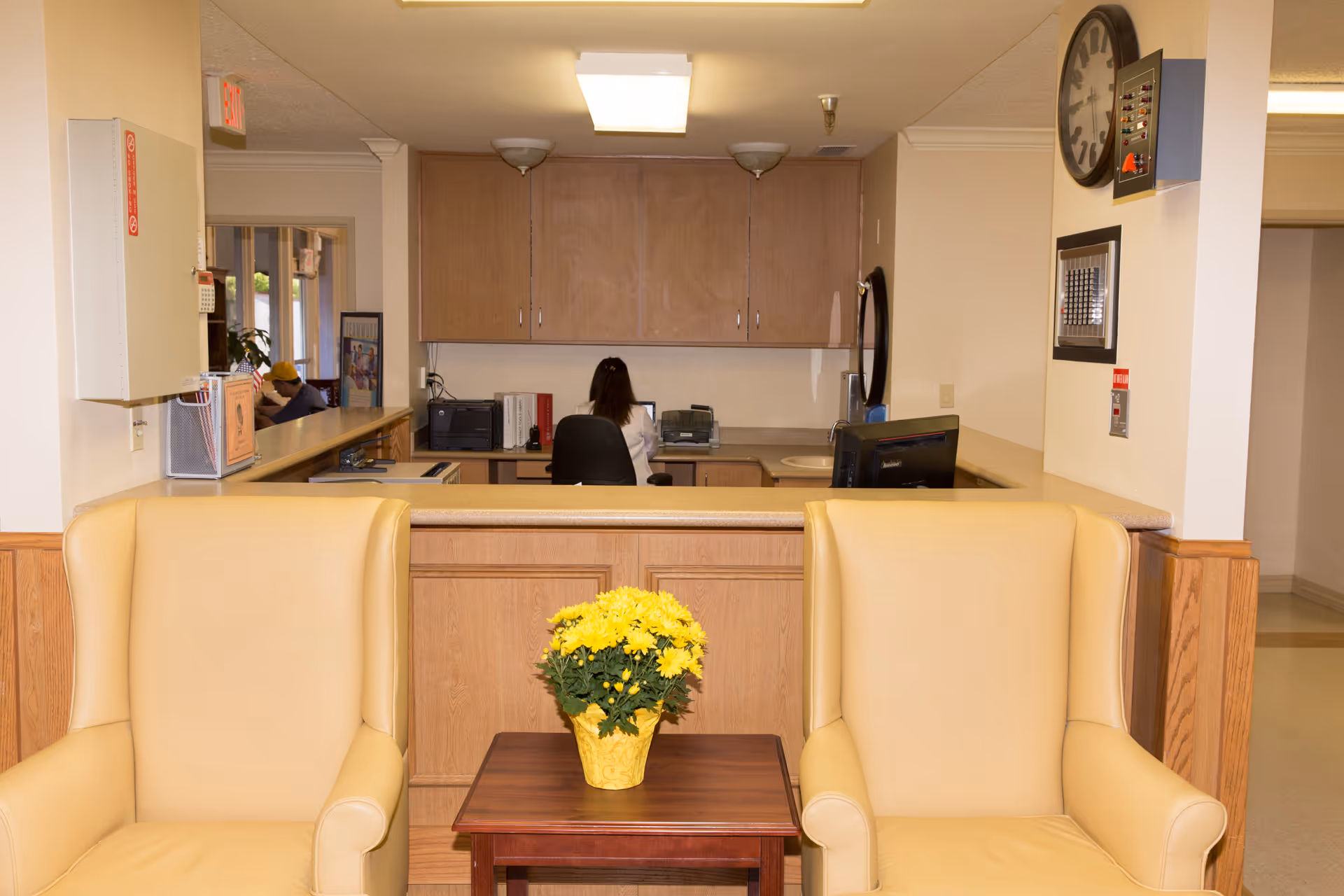 Reception area with beige chairs, a flower centerpiece, and staff at a counter.
