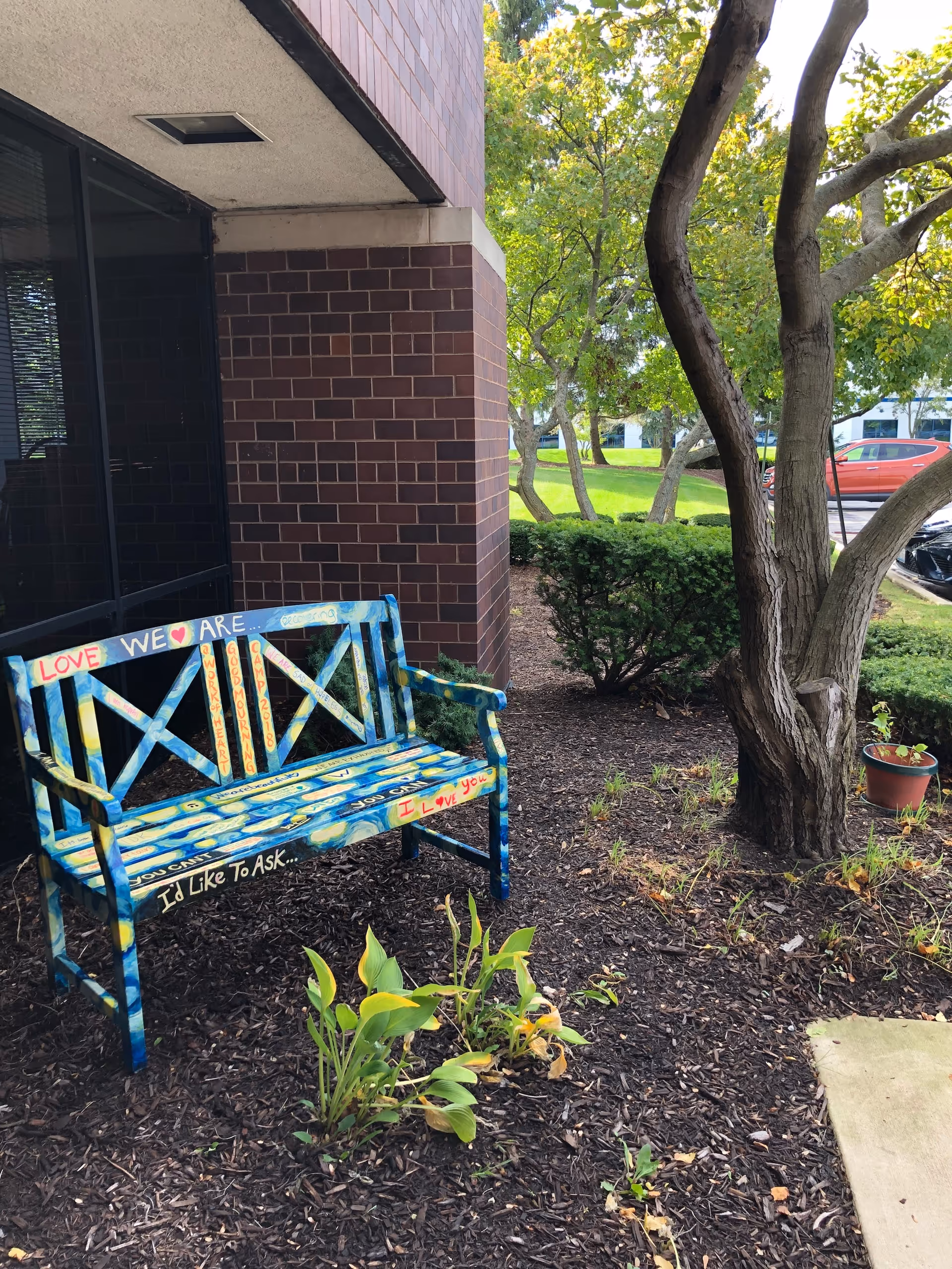 Colorful painted bench outside a brick building with trees and mulch.