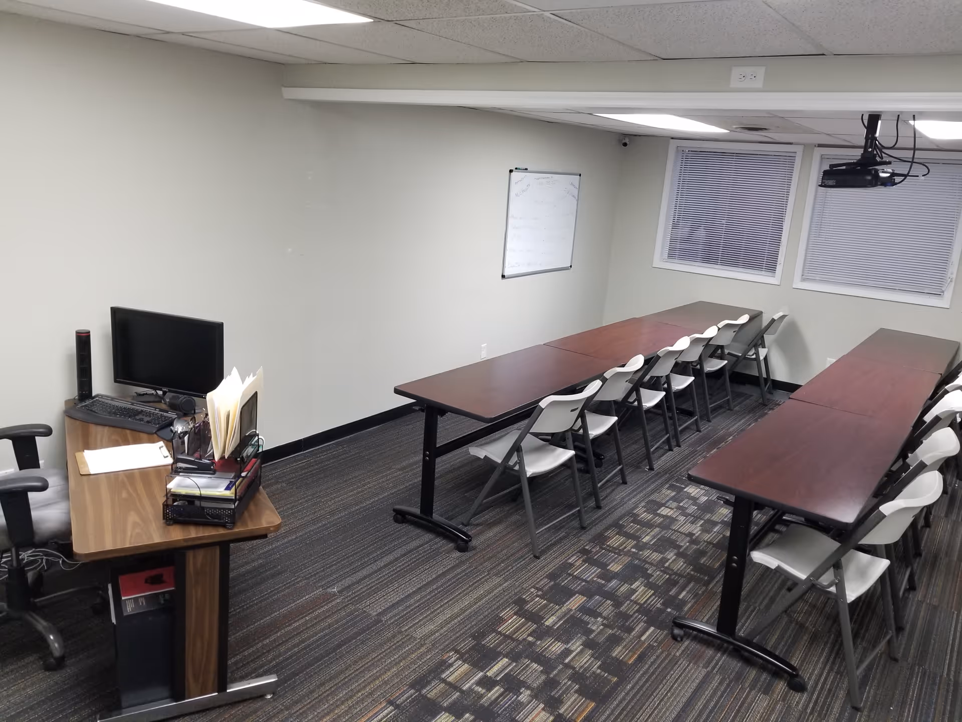 Classroom-style meeting room with long tables, a whiteboard, projector, and stacked chairs.