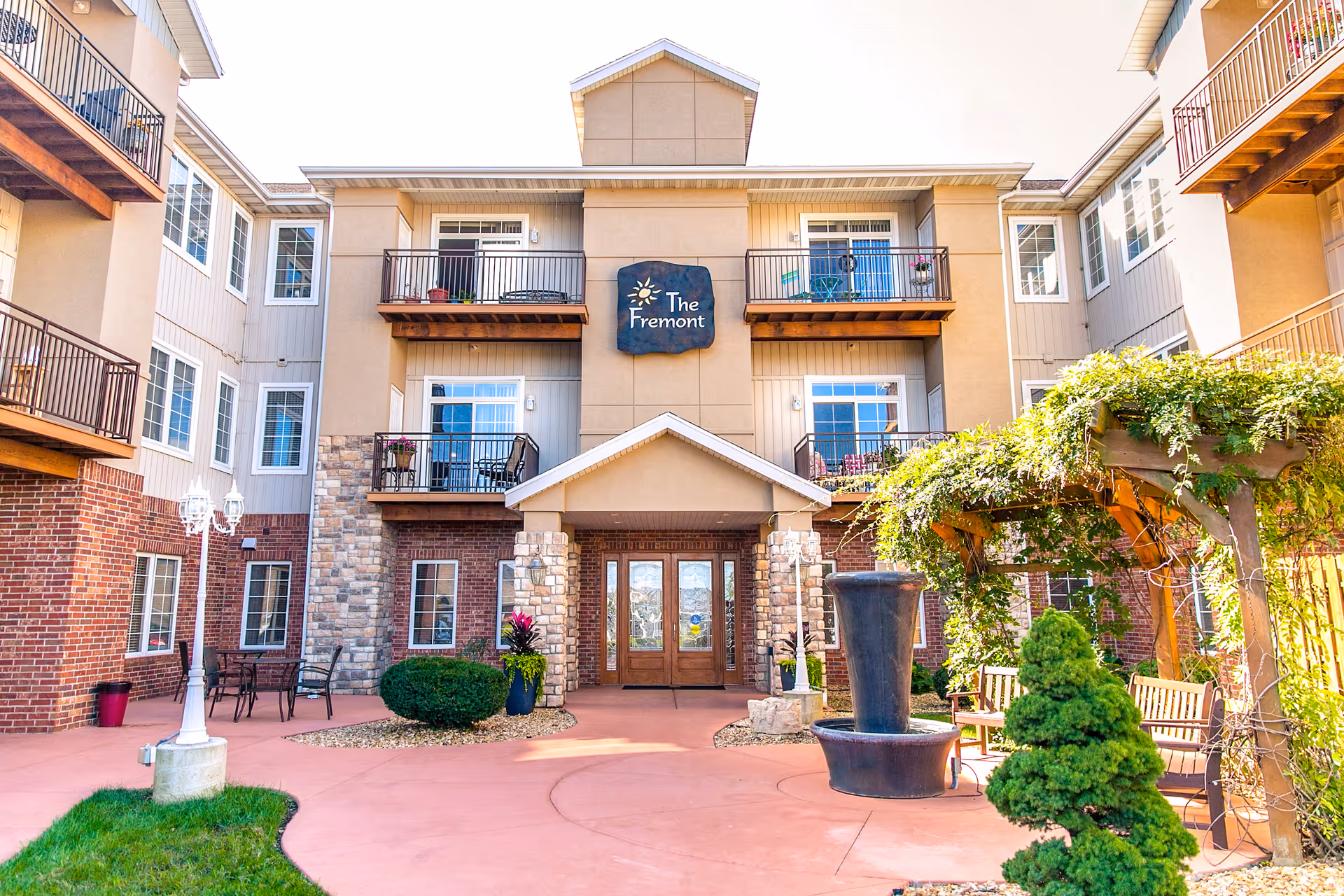 Courtyard view of The Fremont Senior Living building showing the main entrance, balconies, a fountain, and a pergola.