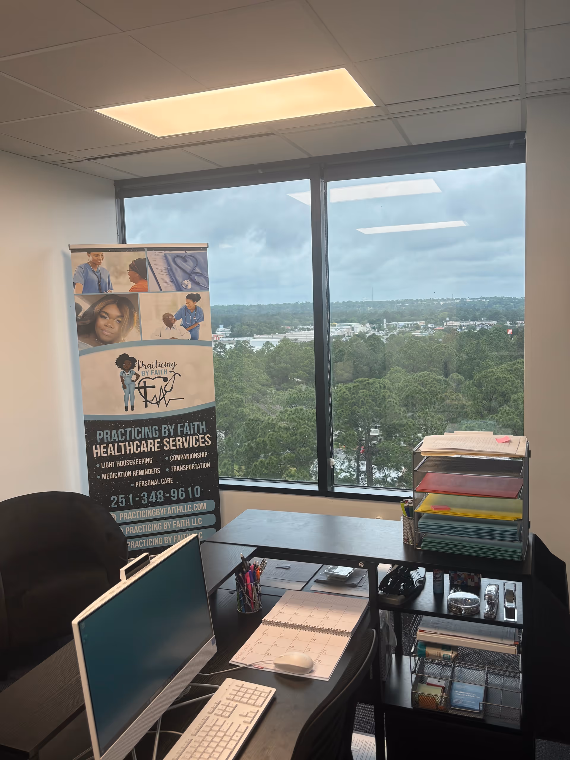 Office desk with computer, calendar, and stacked organizers beside a banner near a large window.