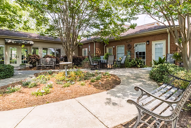 Landscaped courtyard with benches, a round patio table and chairs, trees, and a brick building in the background.
