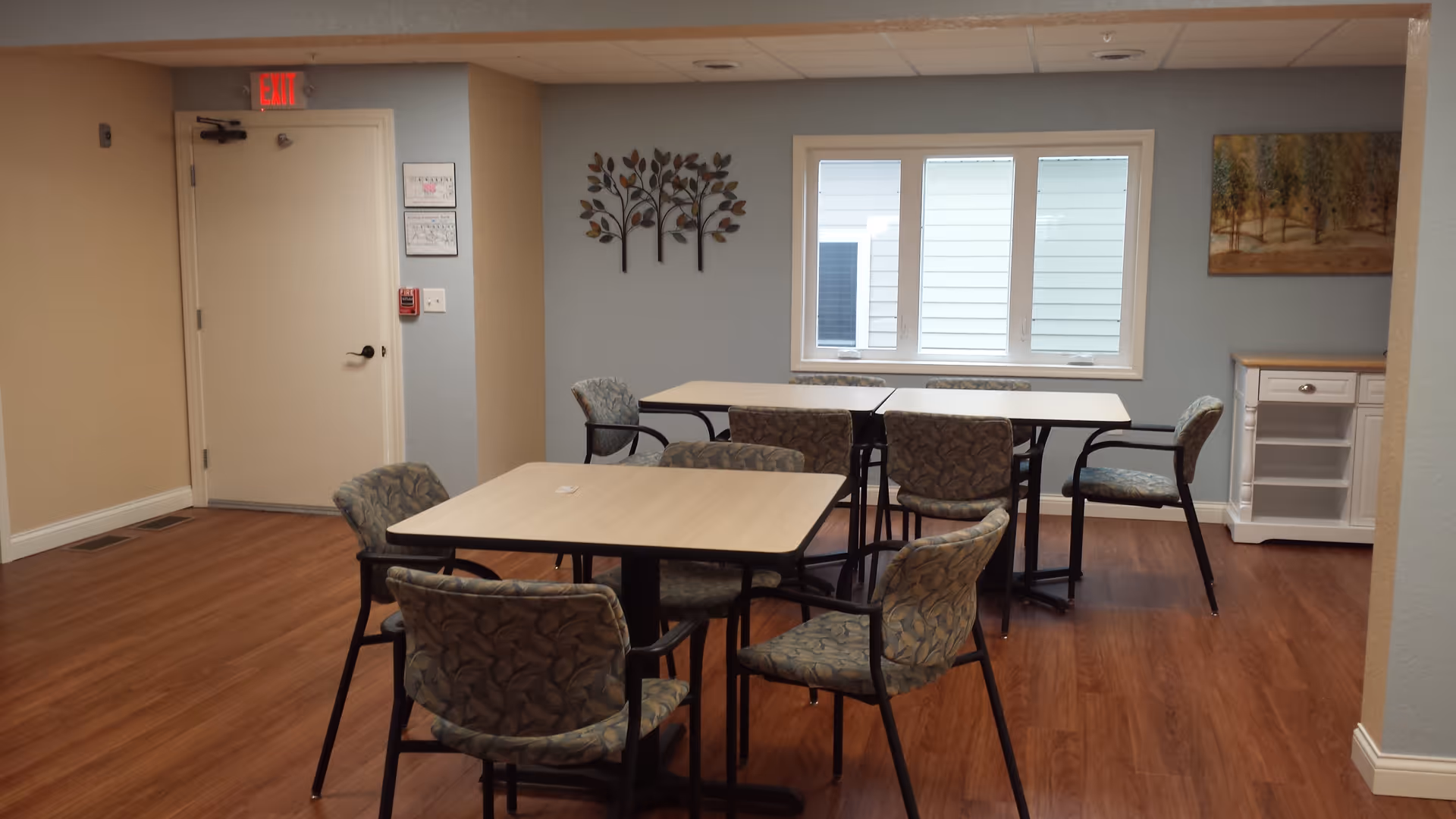 Interior communal dining area with three tables, upholstered chairs, window, and wall art.