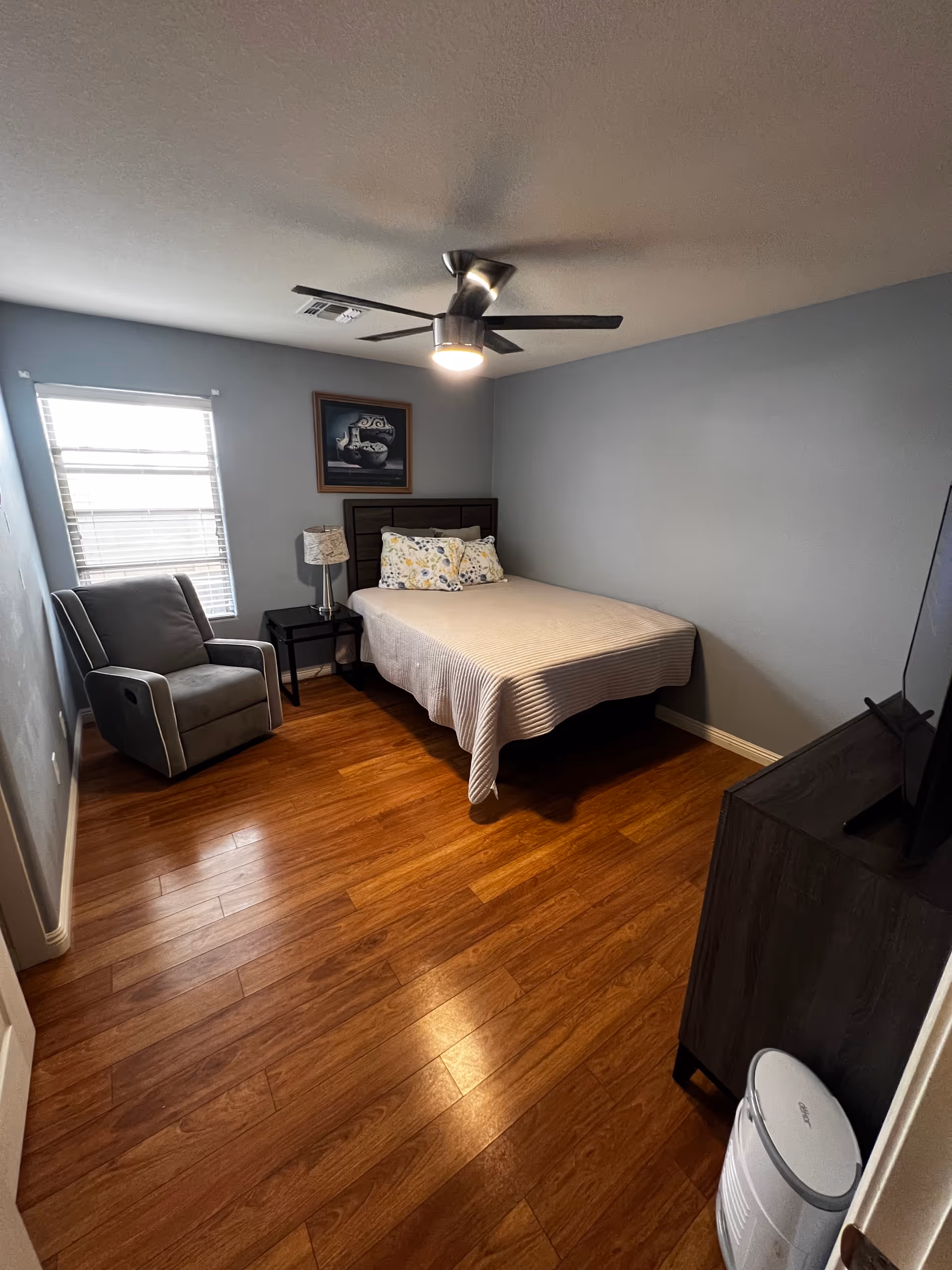 A furnished bedroom with bed, chair, nightstand, dresser, and ceiling fan.