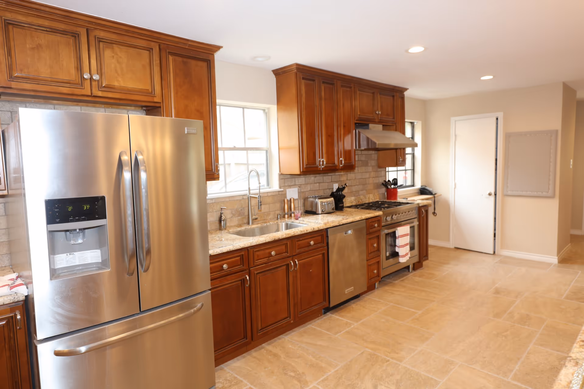 Bright modern kitchen with stainless steel refrigerator, stove, dishwasher, wooden cabinets, granite countertops, and tiled floor.