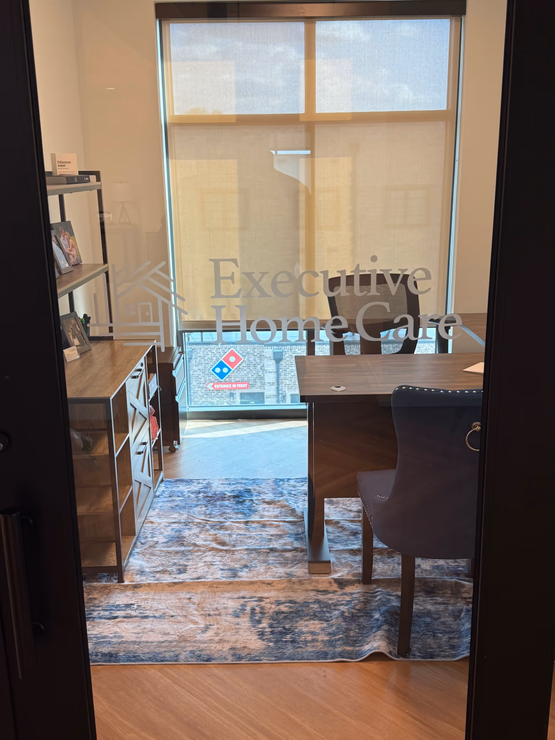 Interior reception area with desk, chair, shelves, rug, and glass door reading Executive Home Care.