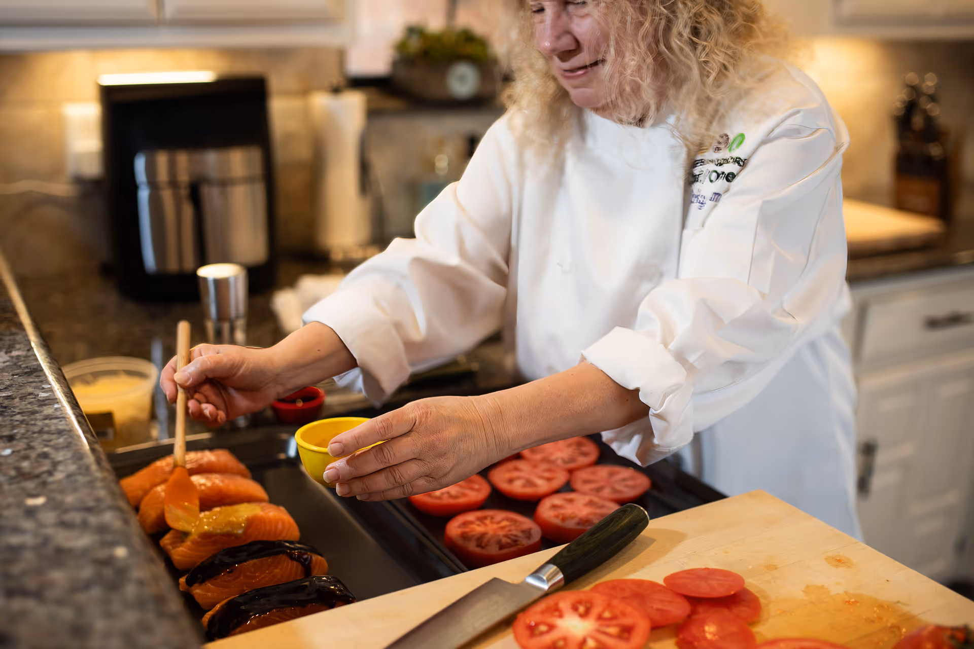 A kitchen staff member prepares salmon and sliced tomatoes on a counter in a facility kitchen.