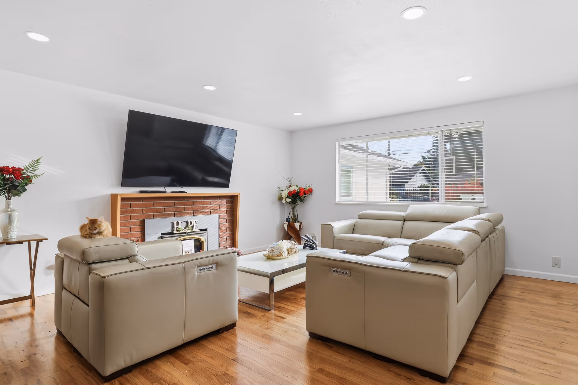 A bright living room with beige leather seating, a brick fireplace, wall-mounted TV, and a cat