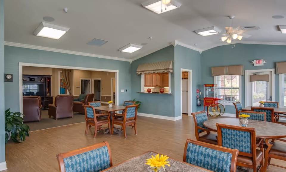 A spacious common area in a senior living facility with several round tables and chairs arranged for dining or socializing. The walls are painted light blue with white trim, and there are large windows letting in natural light. A popcorn machine is visible near the windows, and a doorway leads to a cozy living room area with leather chairs and a television. Ceiling fans and lights are mounted on the ceiling.