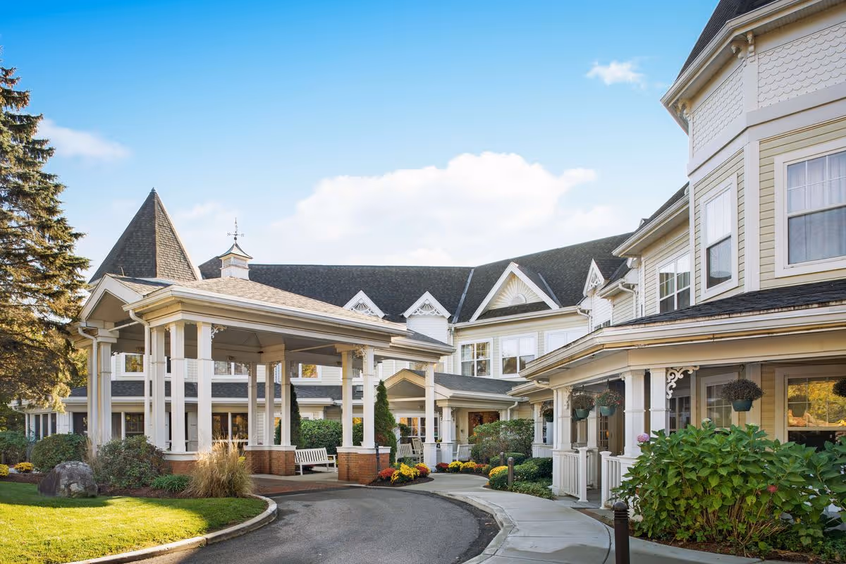 Front exterior of a large Victorian-style senior living facility with a covered porte-cochère, landscaped grounds, and a circular driveway.