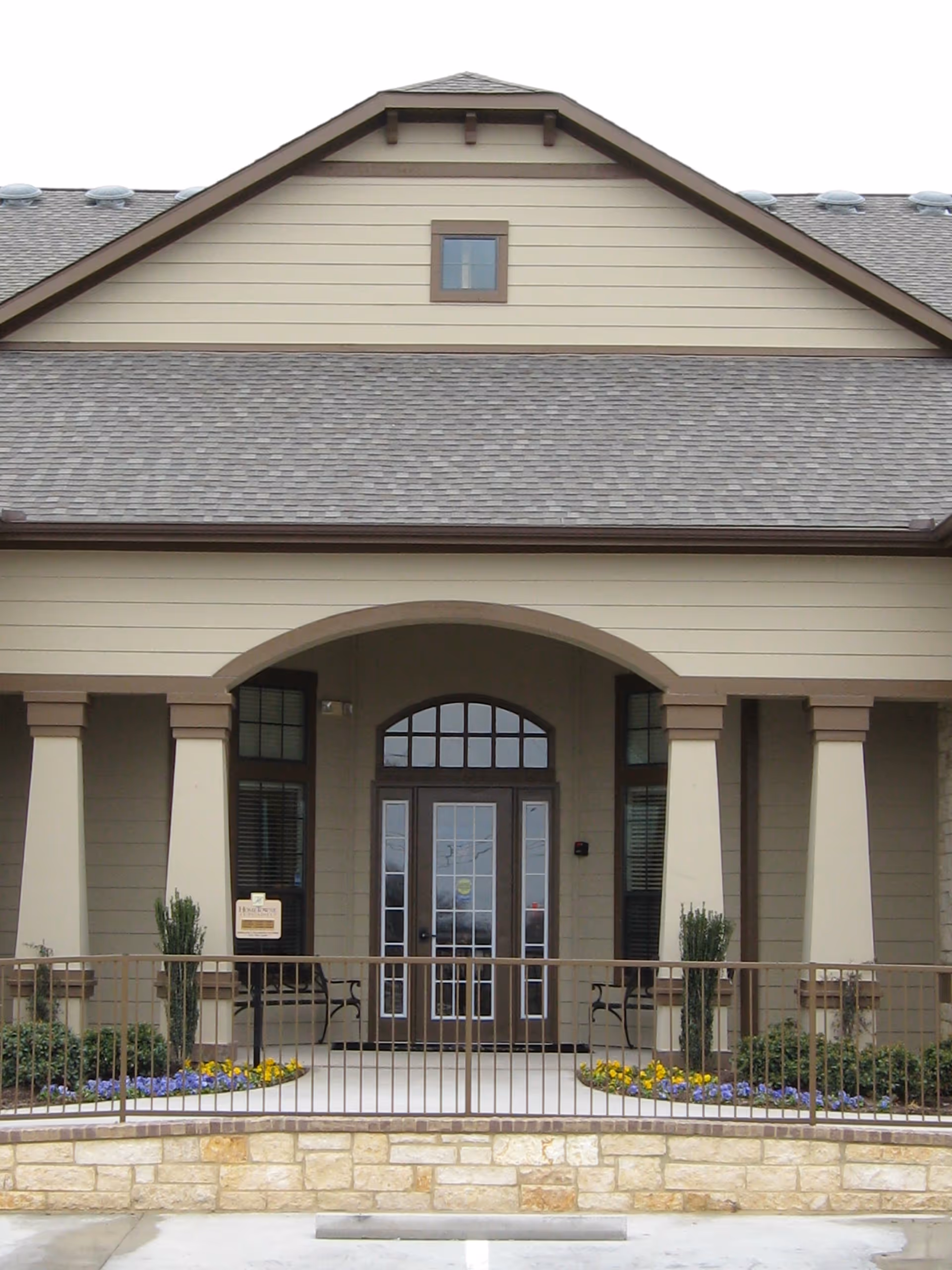 Front entrance facade with an arched porch, columns, double glass doors, and small landscaped flower beds.