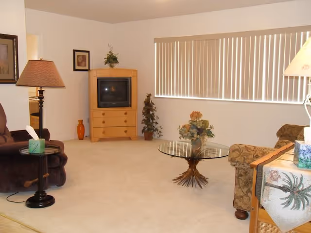 Cozy living room in a senior facility with TV, glass coffee table, and beige carpet.