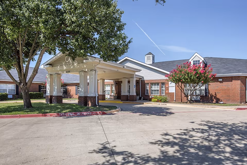 Exterior view of Brookdale Farmers Branch senior living facility showing a covered entrance with white columns, brick walls, windows, and surrounding trees and landscaping under a clear blue sky.