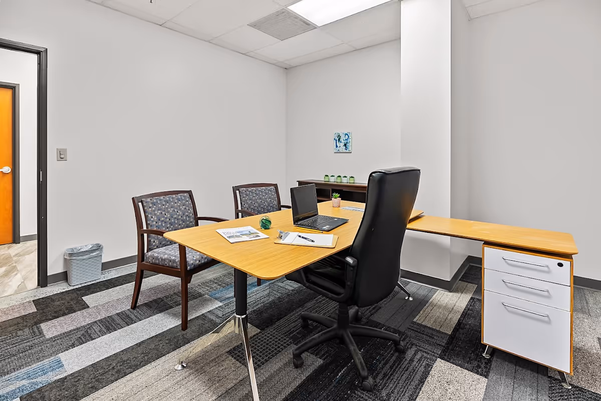 An empty office with a wooden desk, black swivel chair, two guest chairs, and laptop.