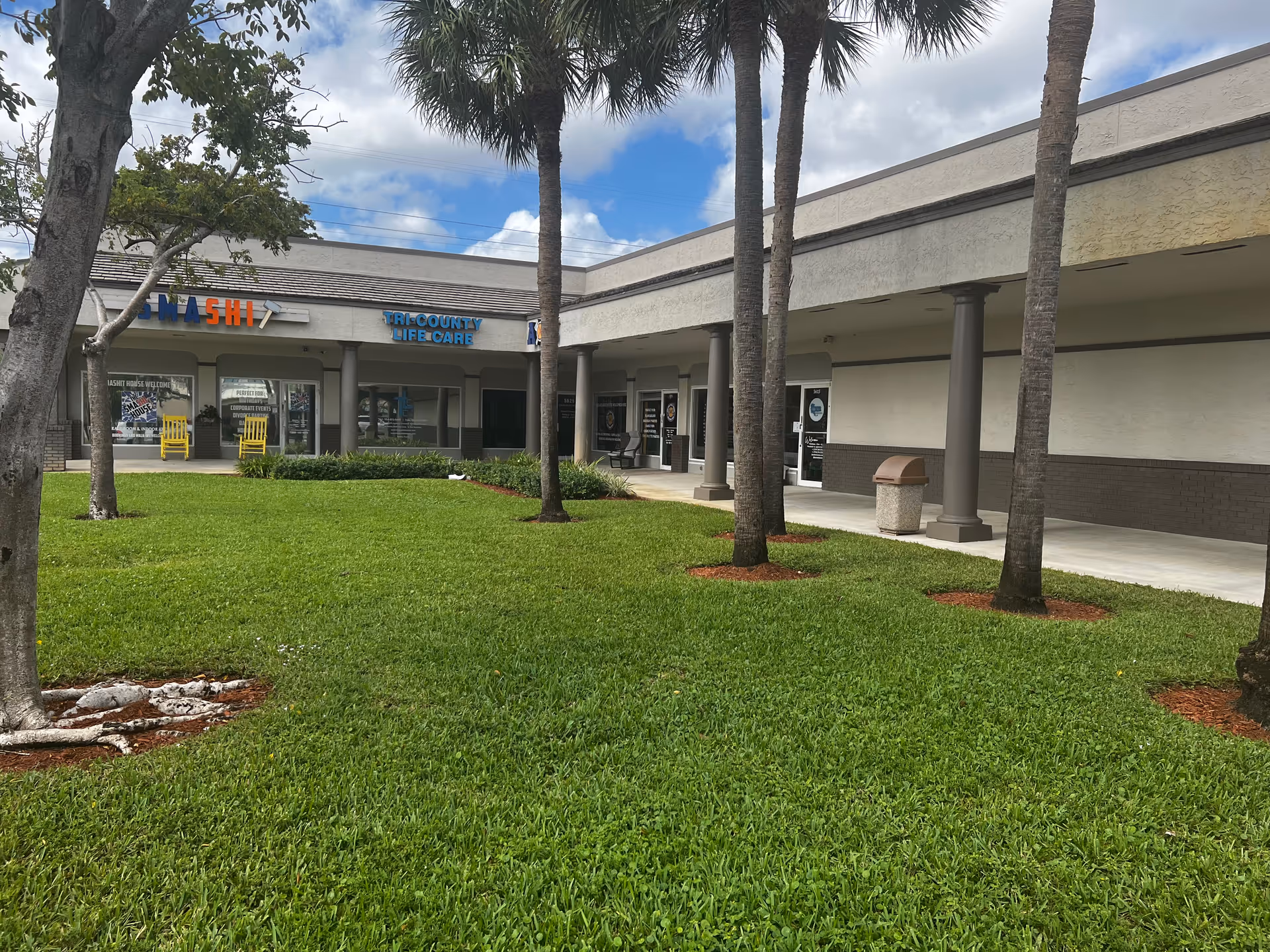 Exterior view of Tri-County Life Care facility with palm trees and storefronts.
