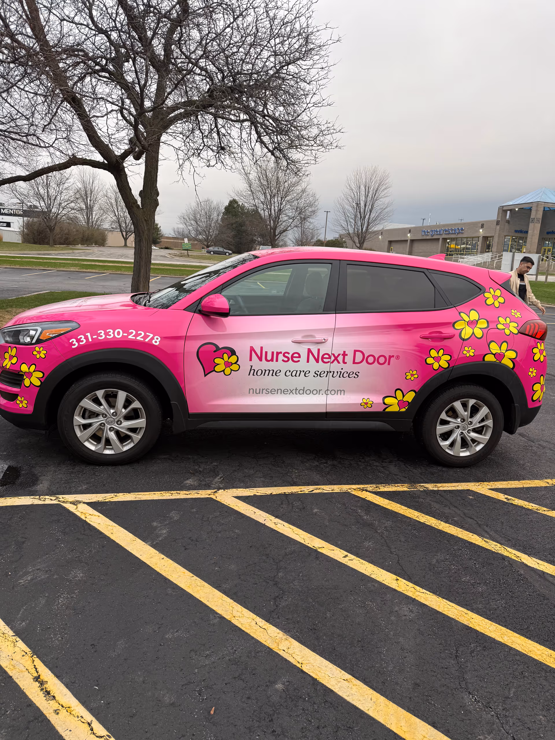 Bright pink Nurse Next Door vehicle with yellow flowers parked in a parking lot.