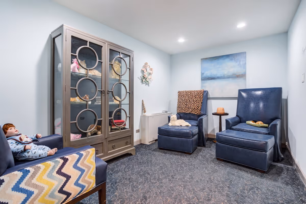 A cozy room with three blue armchairs, one with a colorful zigzag blanket and a doll sitting on it, another with a leopard print blanket and a white stuffed animal, and the third with a small tray holding food. There is a glass-front cabinet displaying various items, a small side table, a wall decoration, and a large abstract painting on the wall. The room has light blue walls and a patterned carpet.