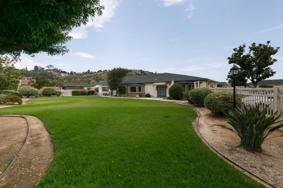 Green lawn and landscaped pathway leading to a single-story senior living building with a white fence and an American flag.